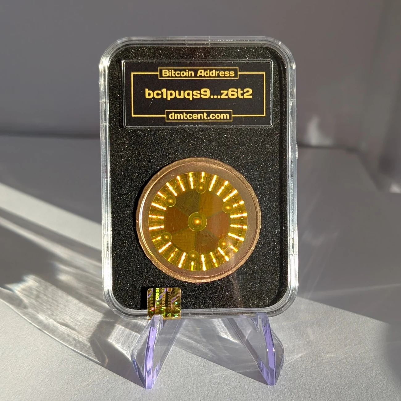 The same coin from the back, where a holographic tamper seal covers the private key. The slab label shows the coin's Bitcoin address in truncated form and the address dmtcent.com.