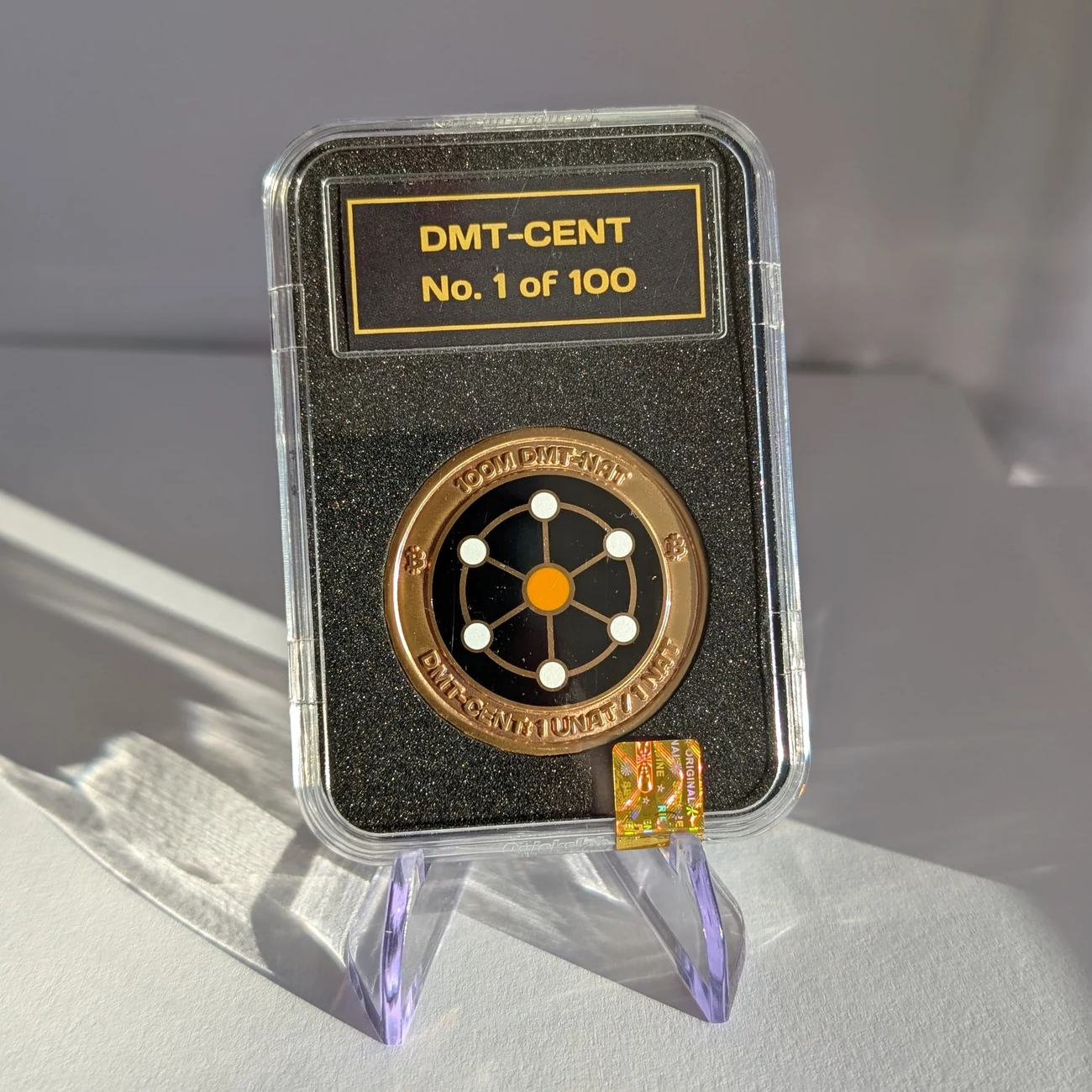 DMT-CENT #1 sealed in its slab, front: a brass coin with the hard-enamel DMT-NAT wheel logo, reading 100M DMT-NAT and DMT-CENT: 1 UNAT / 1 NAT around the rim. The slab label reads DMT-CENT No. 1 of 100.